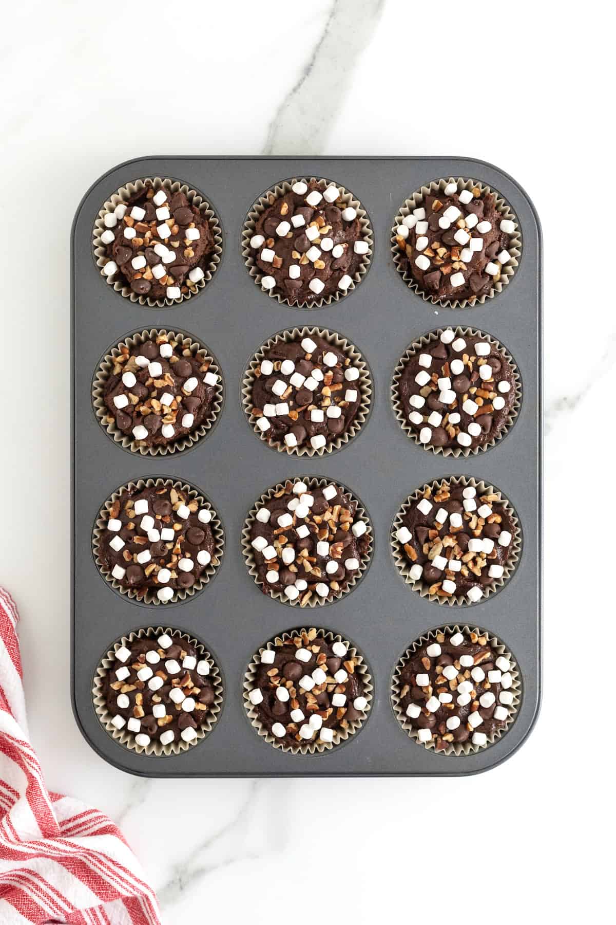 Chocolate brownie cupcakes batter in a muffin tin garnished with mini marshmallows, chocolate chips, and chopped pecans.