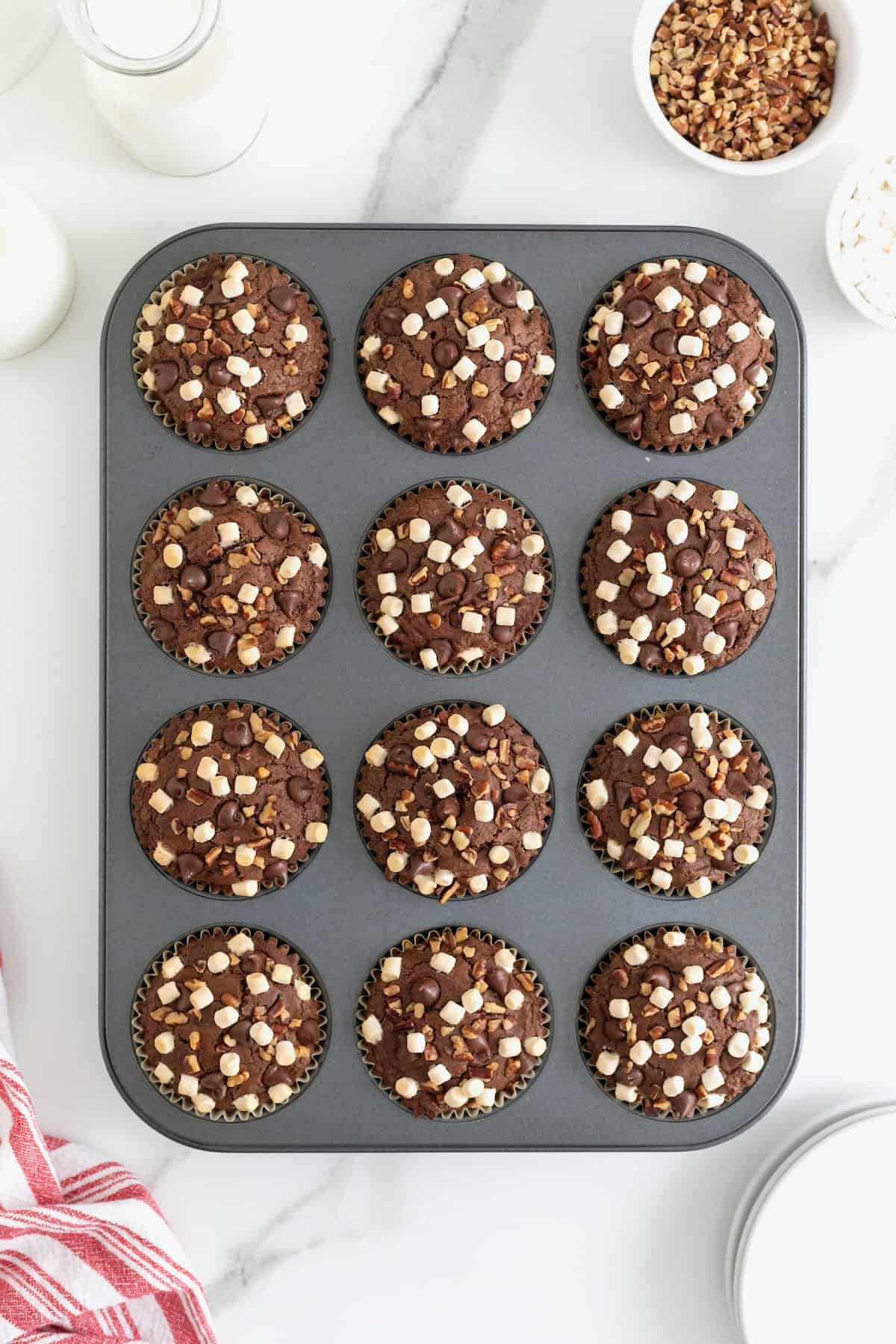 Chocolate brownie cupcakes in a dark aluminum muffin pan garnished with mini marshmallows, chocolate chips, and chopped pecans.