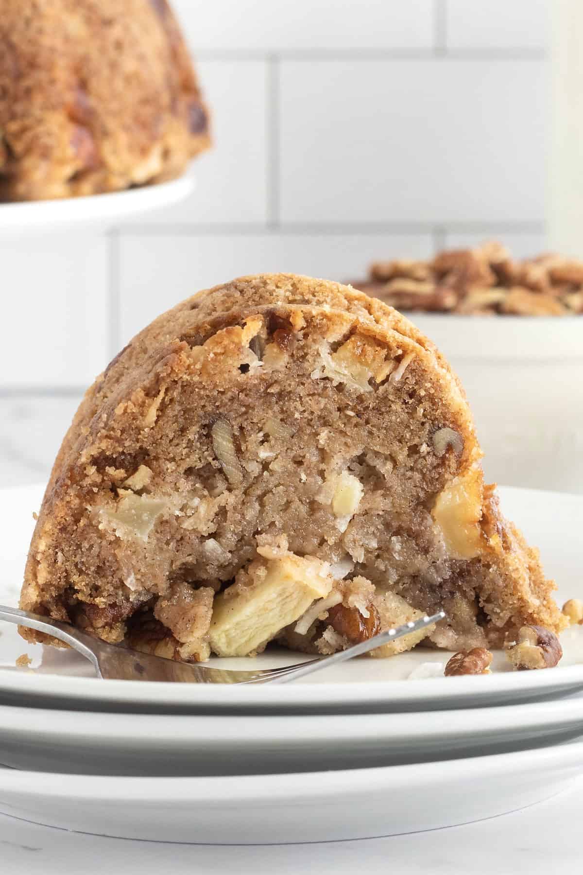 A slice of fresh apple cake with pieces of apple, pecans, and shredded coconut on a stack of three white plates on a white marble counter.