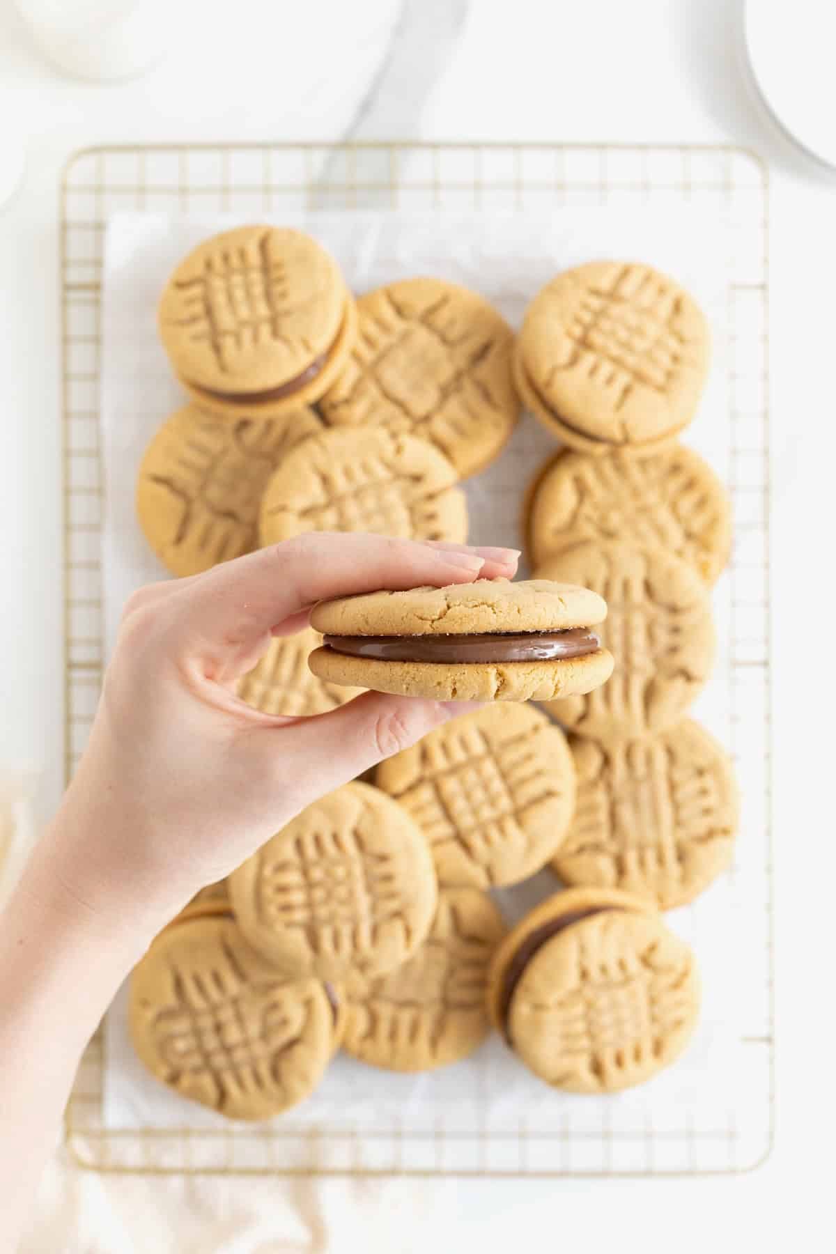 A hand holding a peanut butter sandwich cookie over a pile of cookies on a parchment lined wire cooling rack.