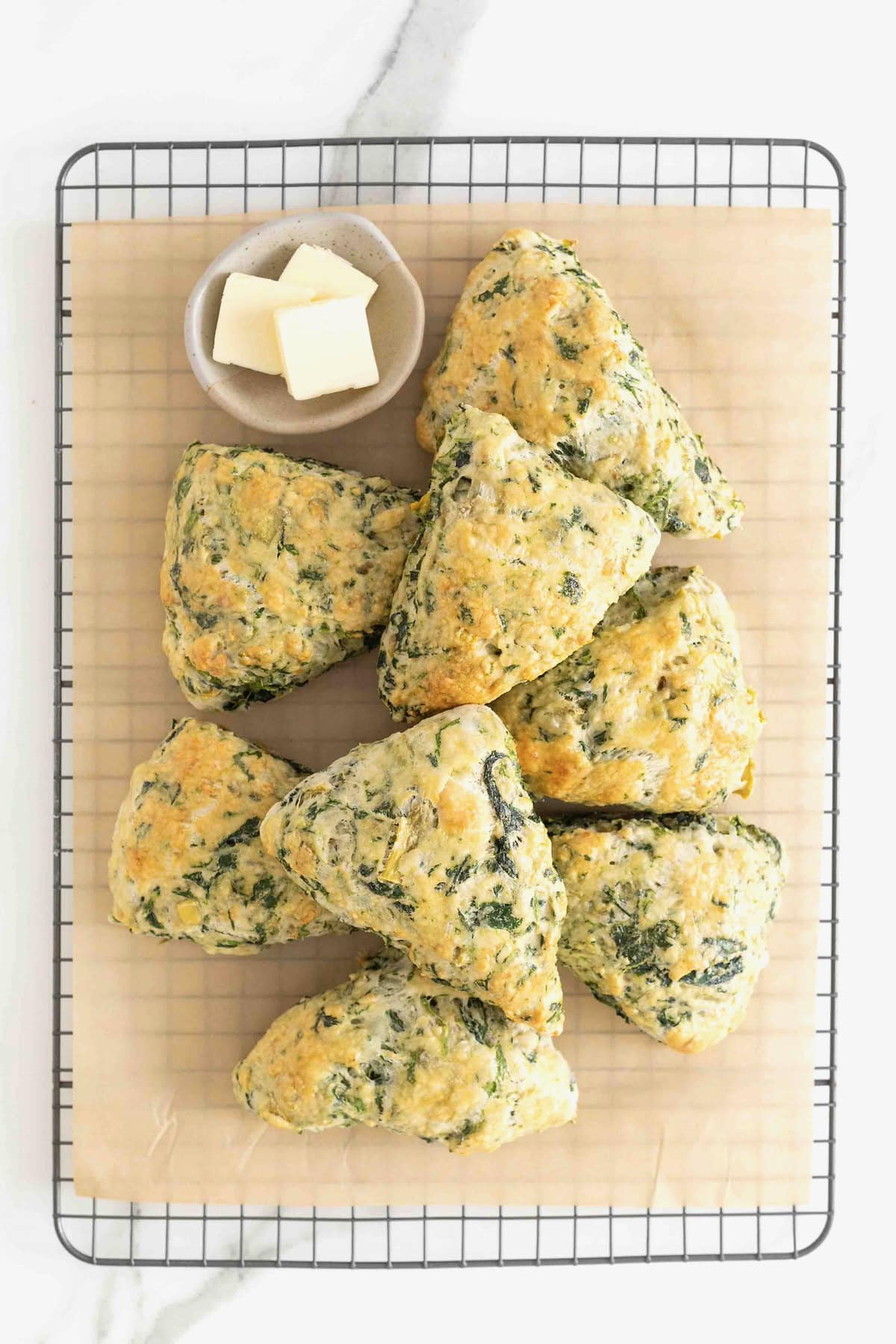 Eight spinach artichoke scones piled on a parchment lined aluminum baking sheet. There is a small dish of butter at the top of the photo.
