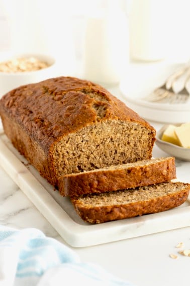 A loaf of banana bread with two slices cut from it on a white marble bread board.