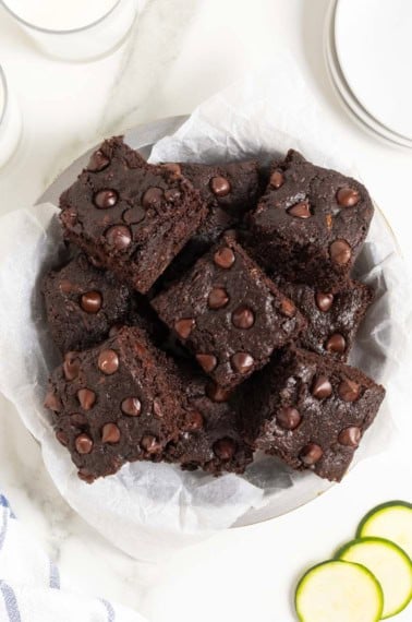 A white plate piled with dark chocolate zucchini brownies on a white marble counter next to several thin slices of zucchini.