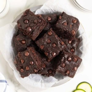 A white plate piled with dark chocolate zucchini brownies on a white marble counter next to several thin slices of zucchini.