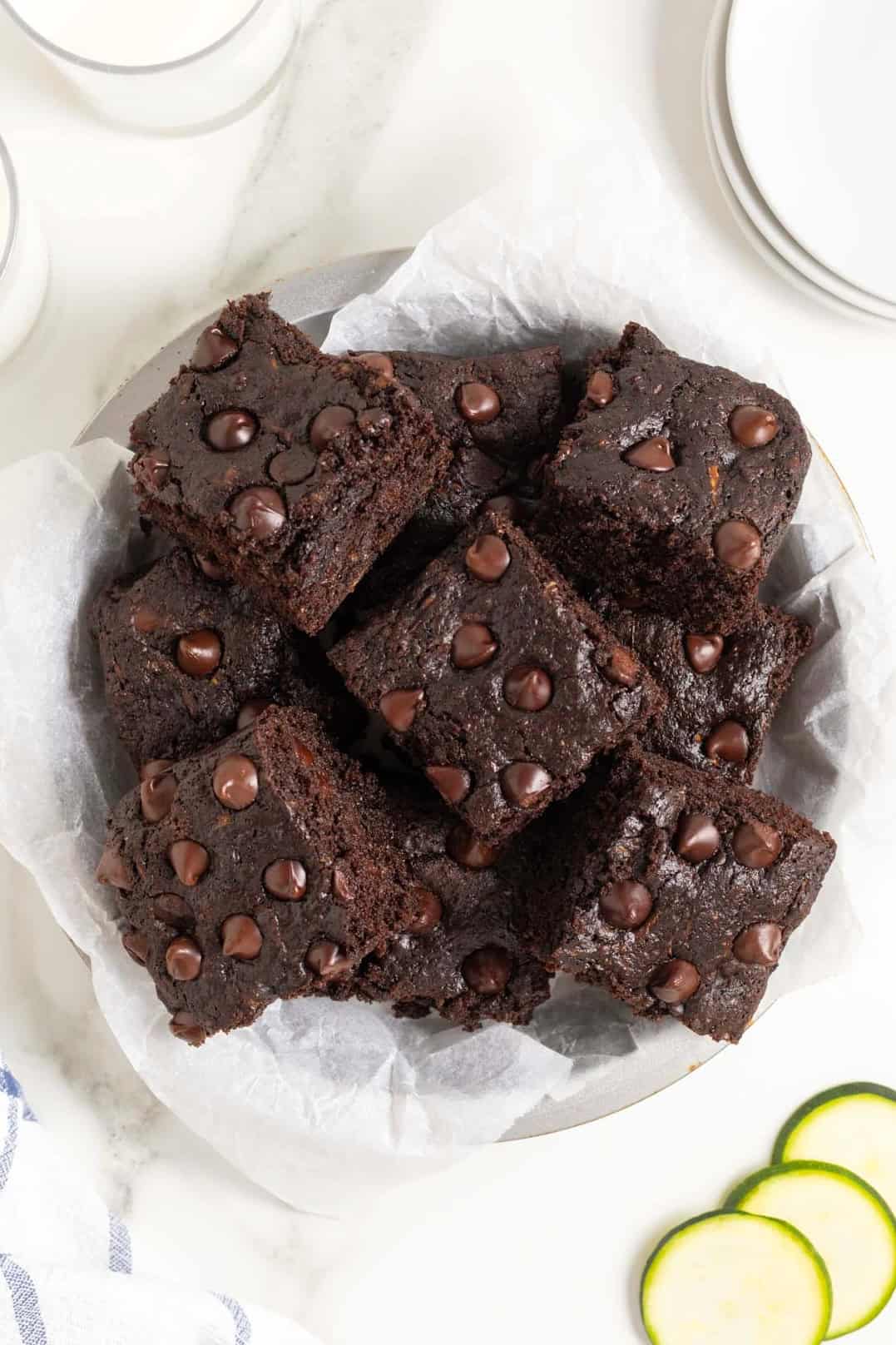 A white plate piled with dark chocolate zucchini brownies on a white marble counter next to several thin slices of zucchini.