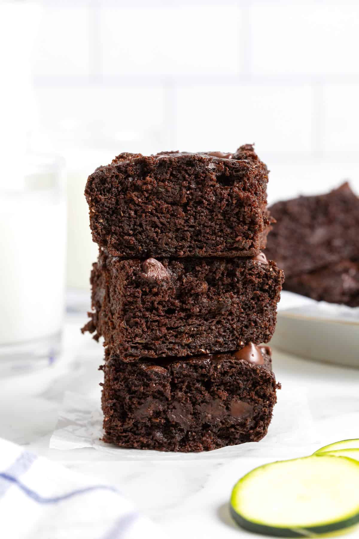 A stack of three zucchini brownies on a white marble counter. A glass of milk is in the background.