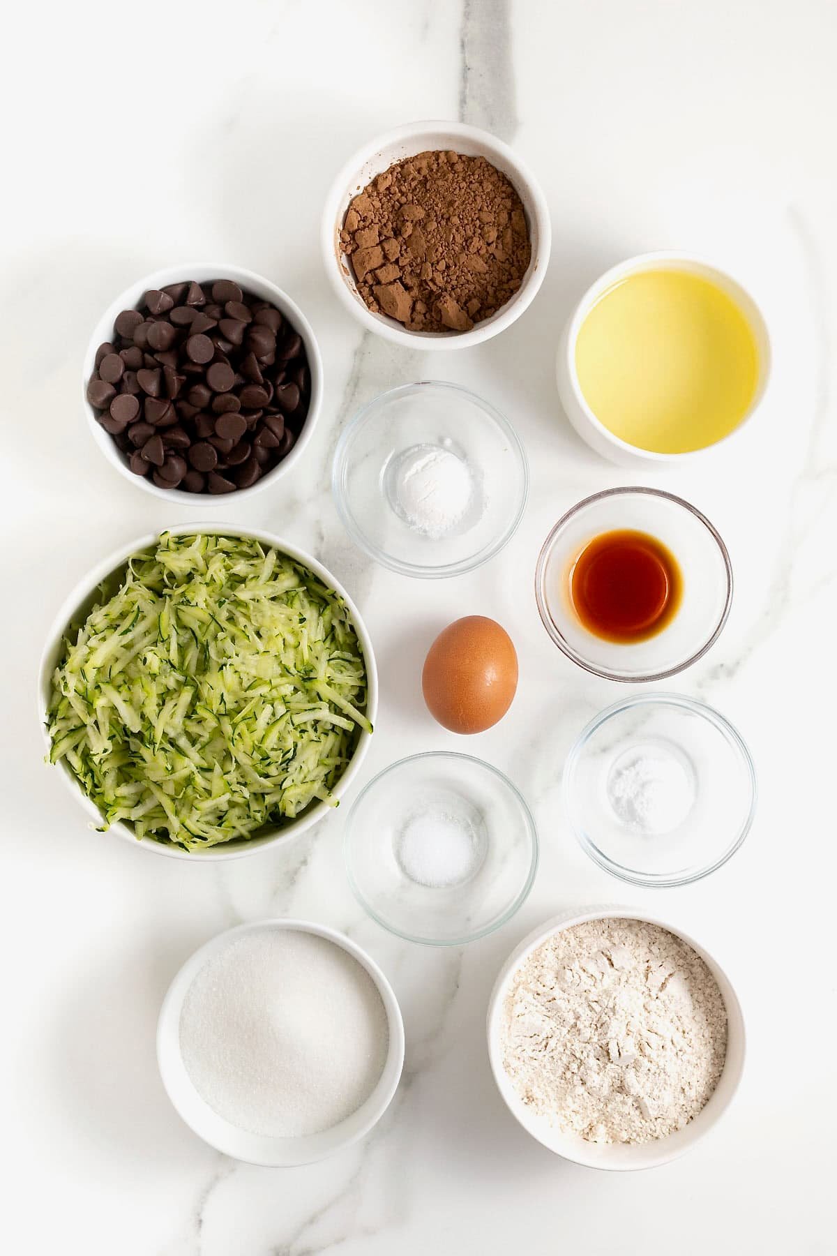 Ingredients to make zucchini brownies in small glass dishes on a white marble counter.