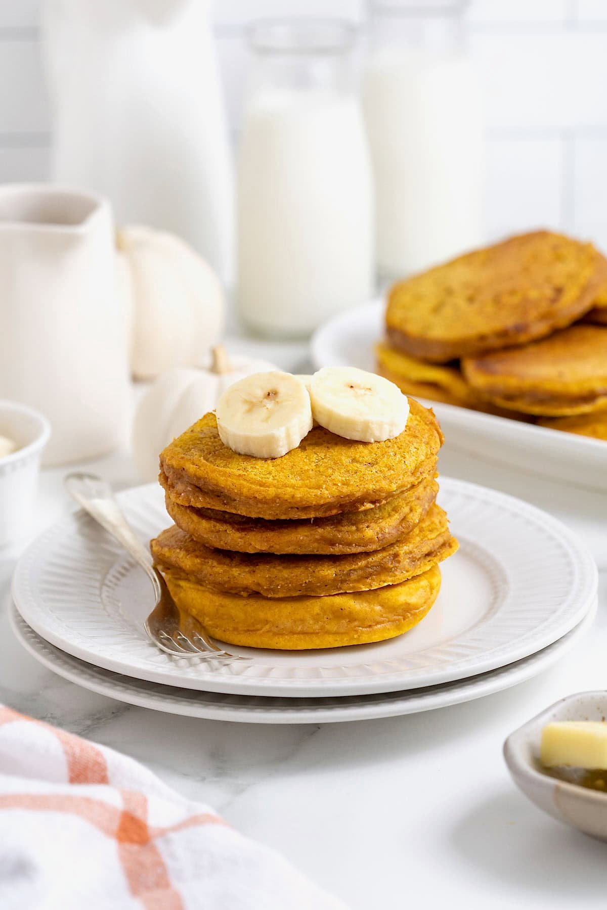 A stack of four pumpkin banana pancakes on a white plate. There are banana slices on top of the pancakes and a fork next to them on the plate.