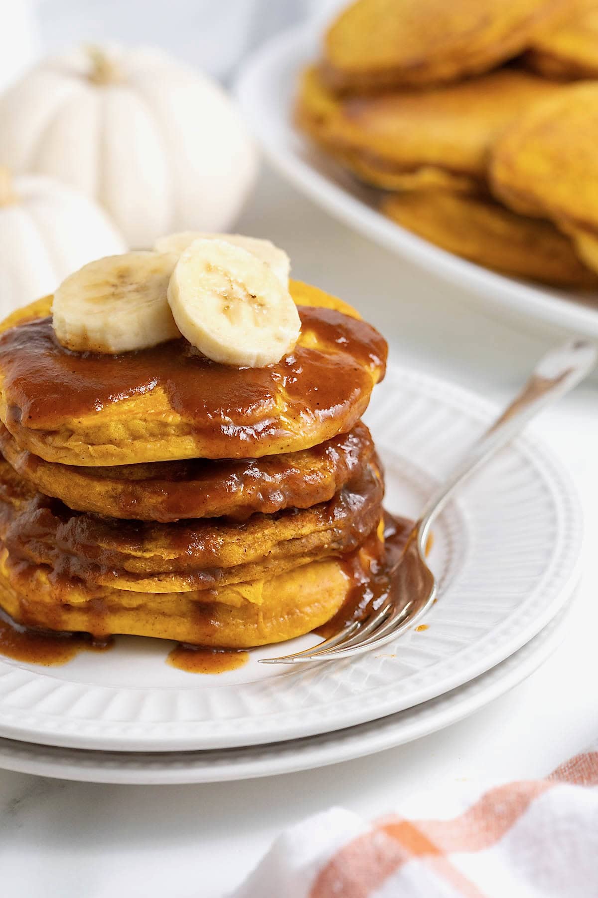 A stack of four banana pumpkin pancakes topped with syrup on a white plate. There are slices of banana on the pancakes and a fork next to them on the plate.