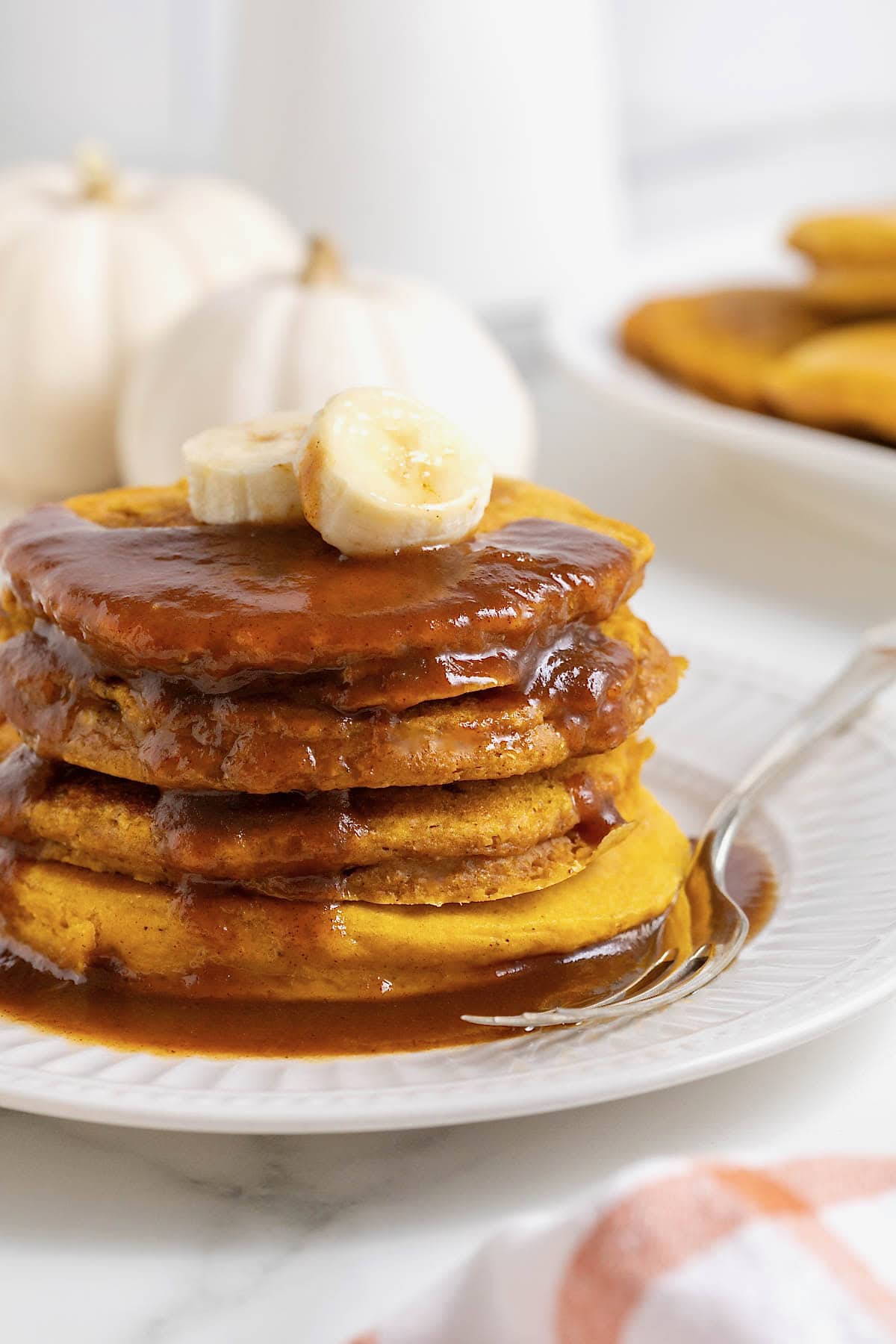 A stack of four banana pumpkin pancakes topped with syrup on a white plate. There are slices of banana on the pancakes and a fork next to them on the plate.
