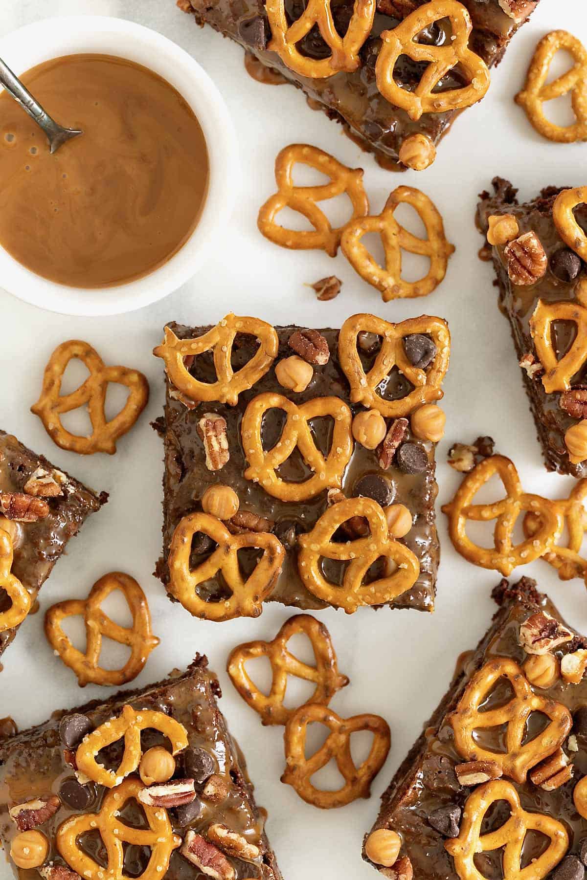 A brownie topped with caramel bits, chocolate chips and pretzels on a white surface surrounded by pretzels and a small white dish of caramel sauce.
