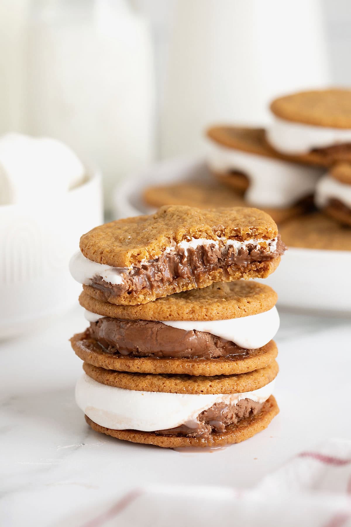 A stack of three s'mores ice cream sandwiches on a white marble surface. The top ice cream sandwich has a bite out of it.