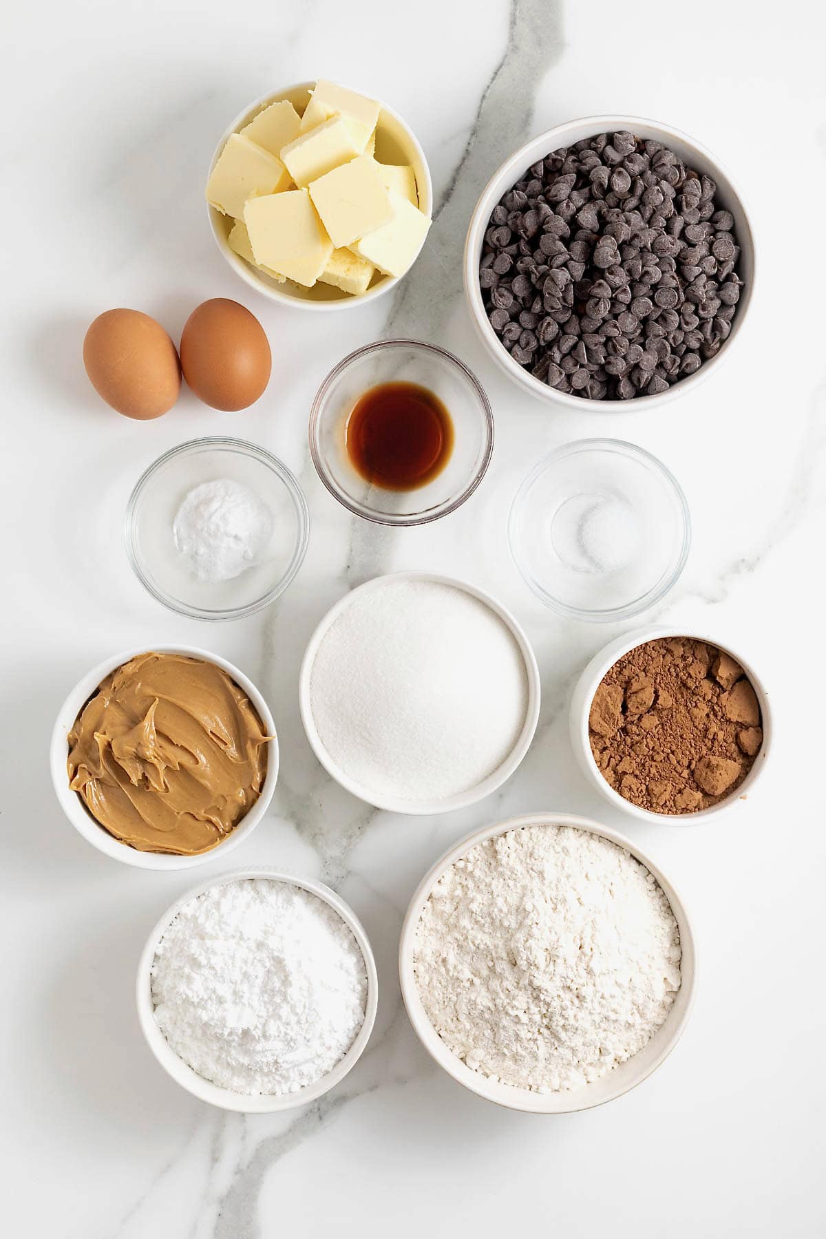 Ingredients to make peanut butter chocolate layered cookie cake in small glass dishes on a white marble counter.