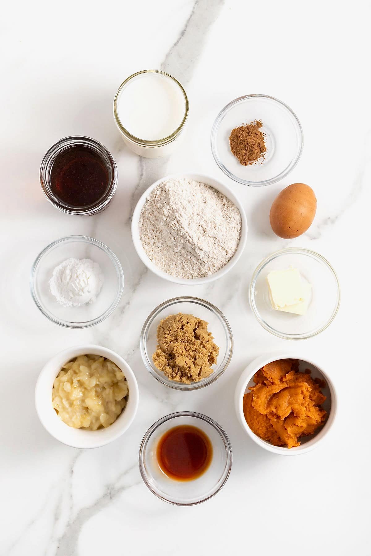 Ingredients to make pumpkin banana pancakes in small glass dishes on a white marble counter.