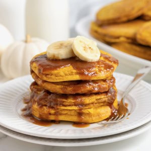 A stack of four banana pumpkin pancakes topped with syrup on a white plate. There are slices of banana on the pancakes and a fork next to them on the plate.