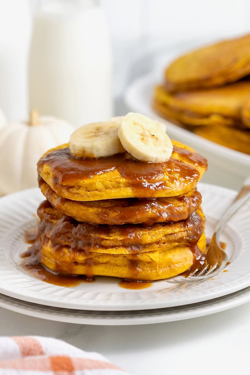 A stack of four banana pumpkin pancakes topped with syrup on a white plate. There are slices of banana on the pancakes and a fork next to them on the plate.