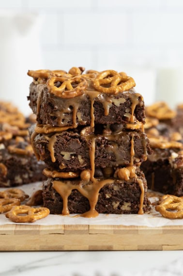 Three brownies in a stack on a white surface. The brownies are dripping with caramel and topped with with pretzels.