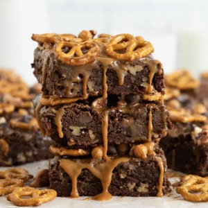 Three brownies in a stack on a white surface. The brownies are dripping with caramel and topped with with pretzels.