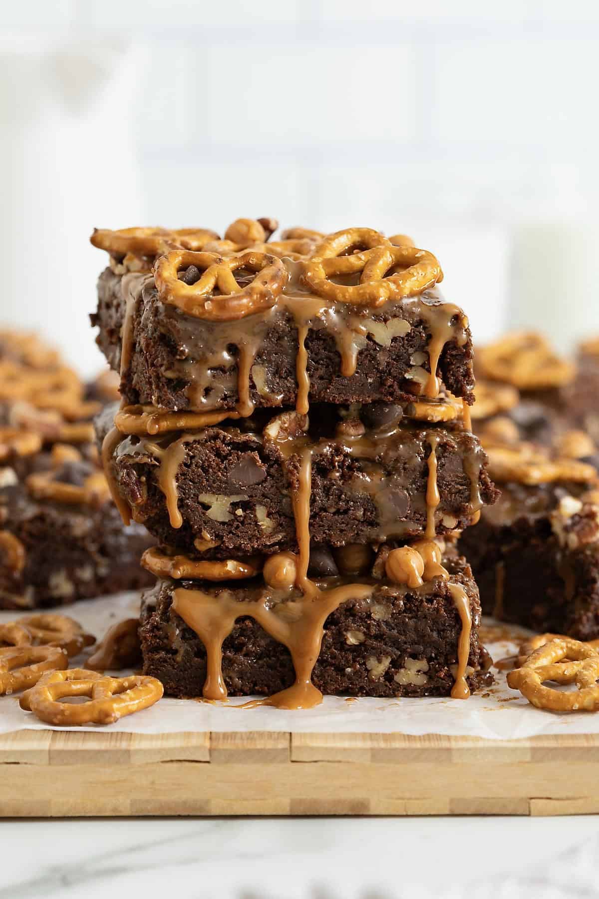 Three brownies in a stack on a white surface. The brownies are dripping with caramel and topped with with pretzels.