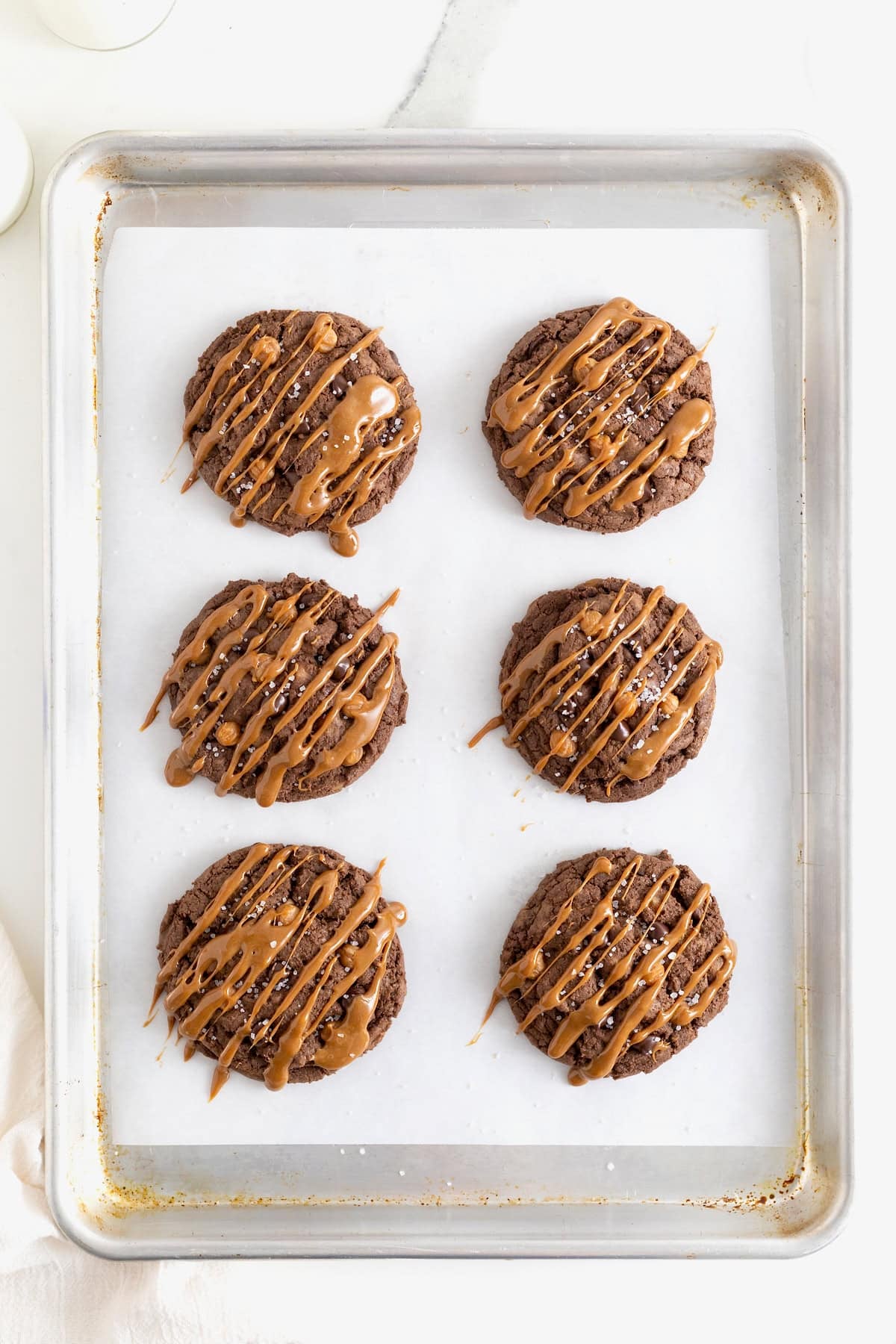 Six dark chocolate cookies drizzled with caramel on a parchment lined baking sheet.