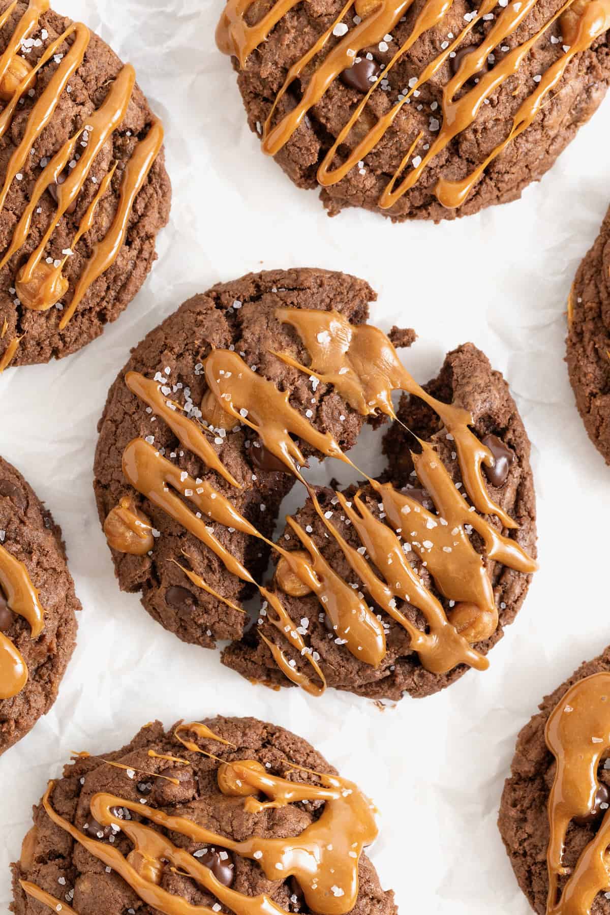 Seven large chocolate cookies with chocolate chips and caramel bits drizzled in caramel sauce on a white surface.