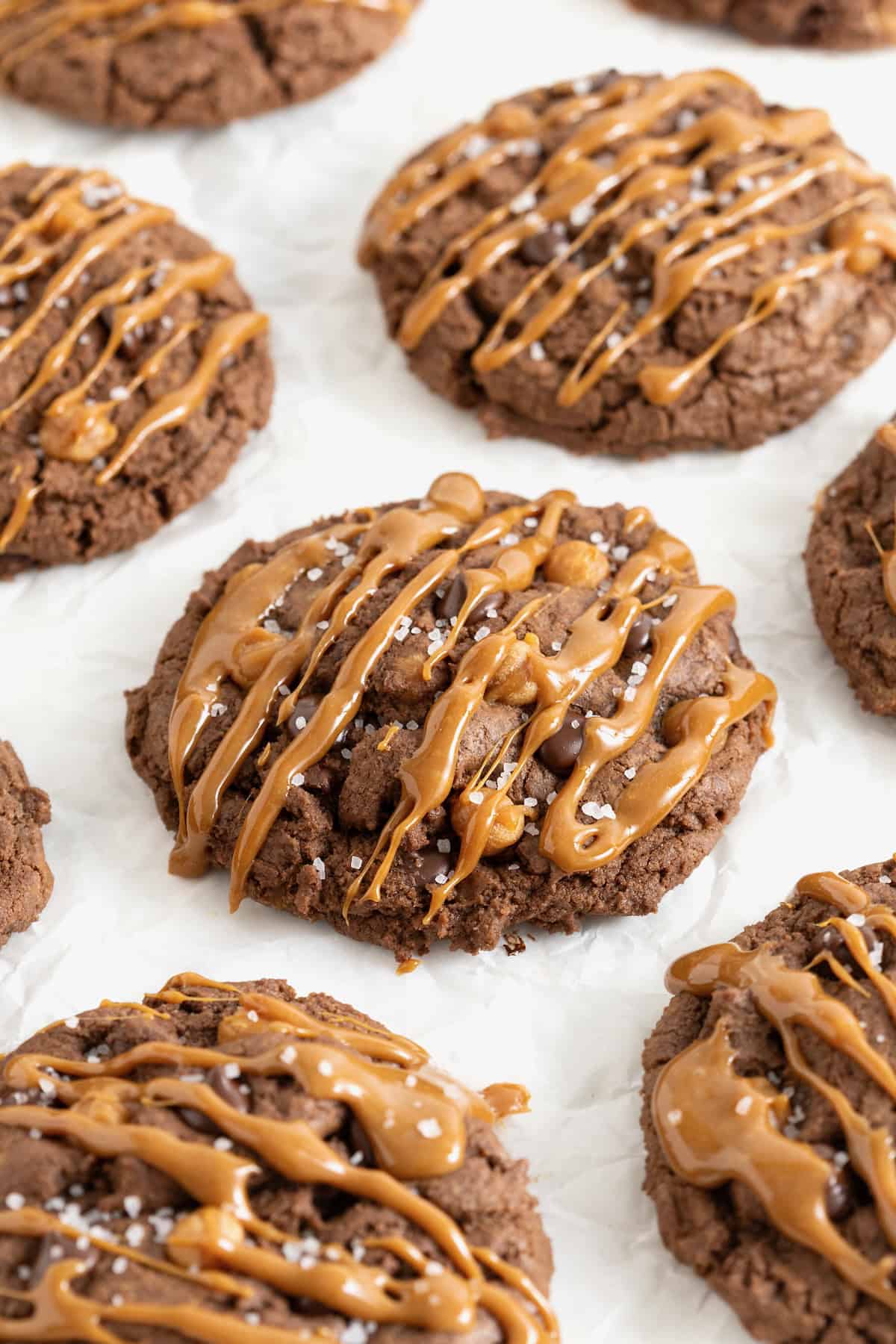 Seven large chocolate cookies with chocolate chips and caramel bits drizzled in caramel sauce on a white surface.