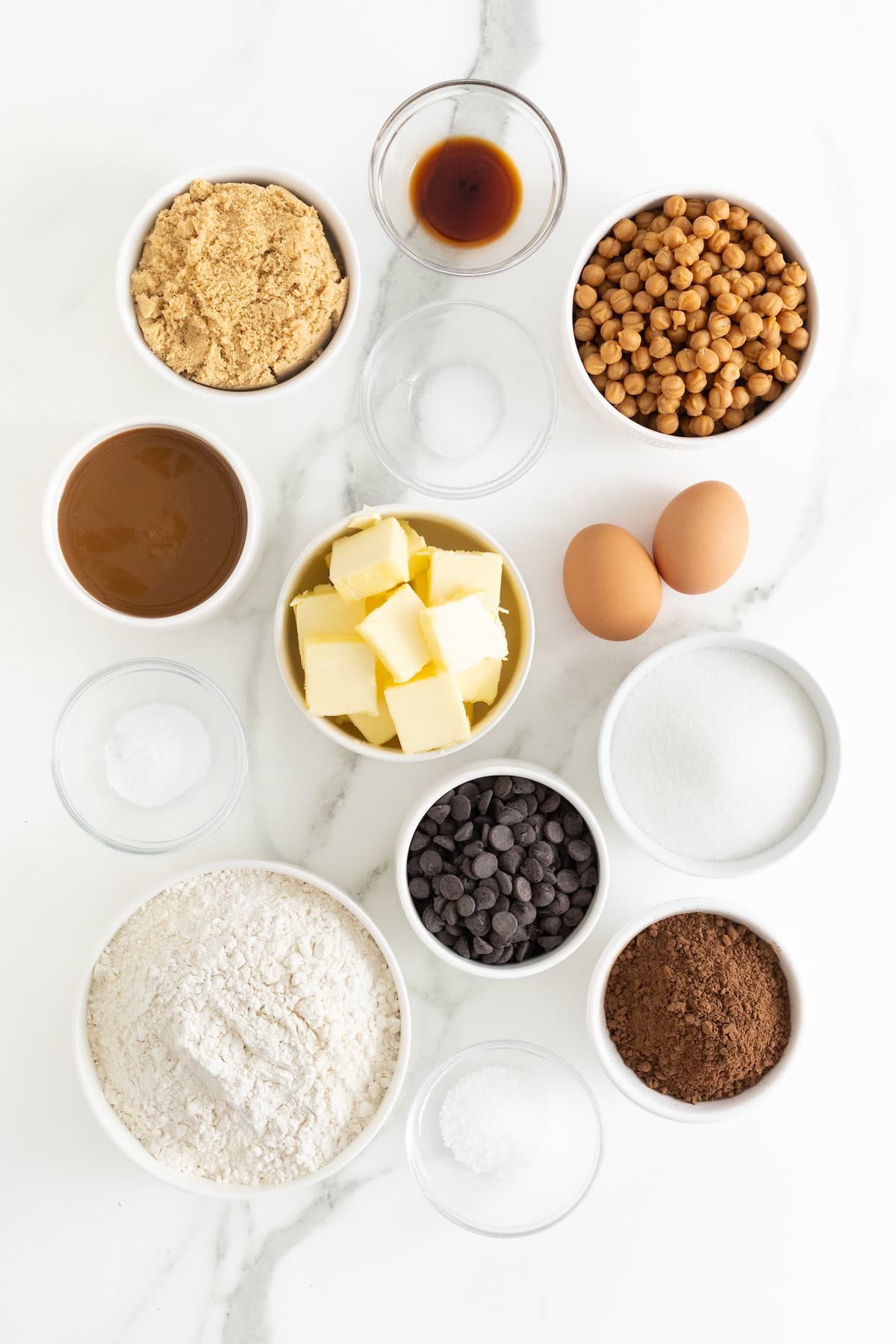 Ingredients to make salted caramel dark chocolate cookies in small glass dishes on a white marble counter.