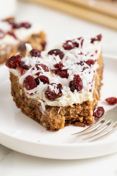 A cookie bar topped with white chocolate, coconut flakes and dried cranberries on a white rimmed plate.