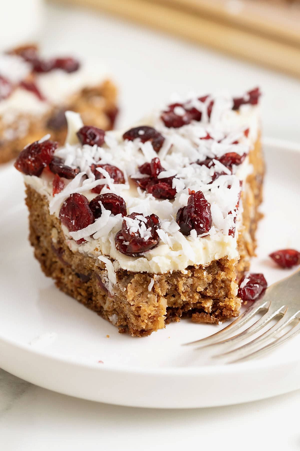 A cookie bar topped with white chocolate, coconut flakes and dried cranberries on a white rimmed plate.
