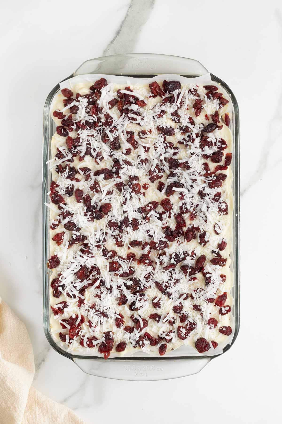 A parchment lined glass baking dish with baked cookie bars topped with melted white chocolate, shredded coconut and dried cranberries.