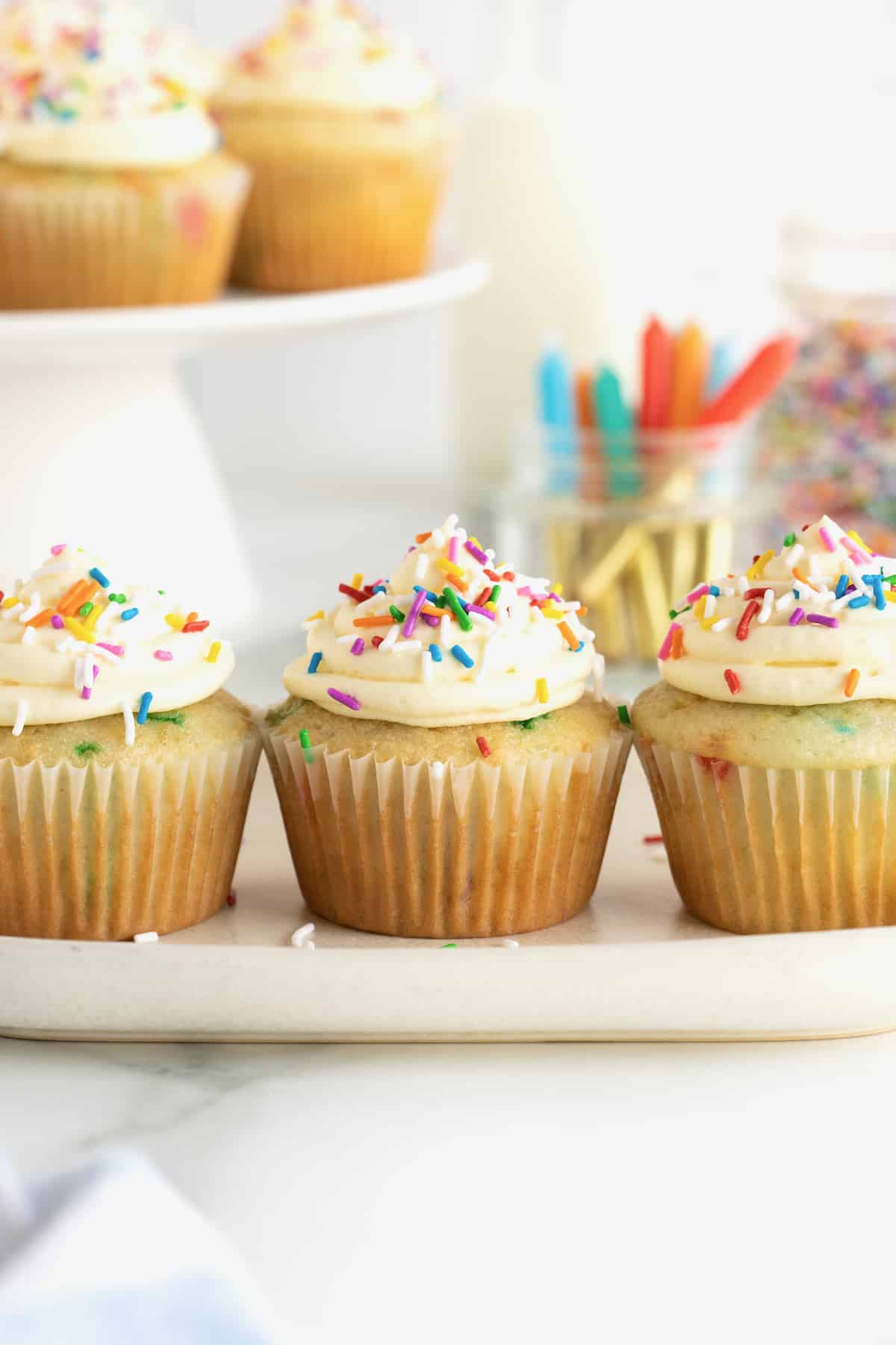 Three vanilla confetti cupcakes with white frosting and colored sprinkles on a white platter.
