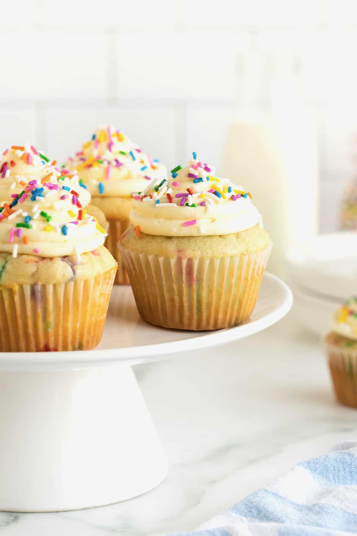 A white porcelain cake stand with four vanilla confetti cupcakes with vanilla frosting and colorful confetti sprinkles.