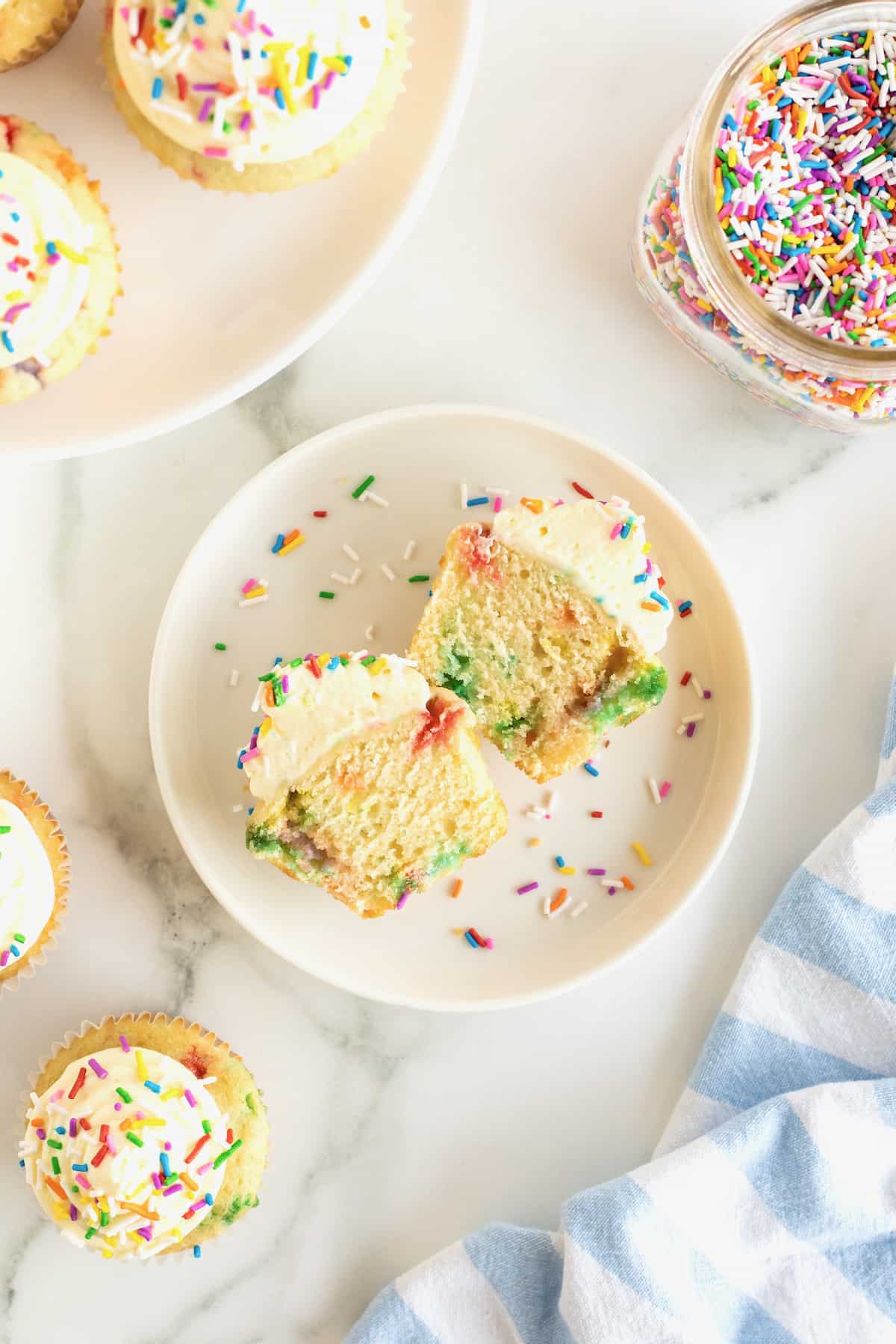 A vanilla confetti cupcake cut in Hal on a white rimmed plate with colorful sprinkles around it.