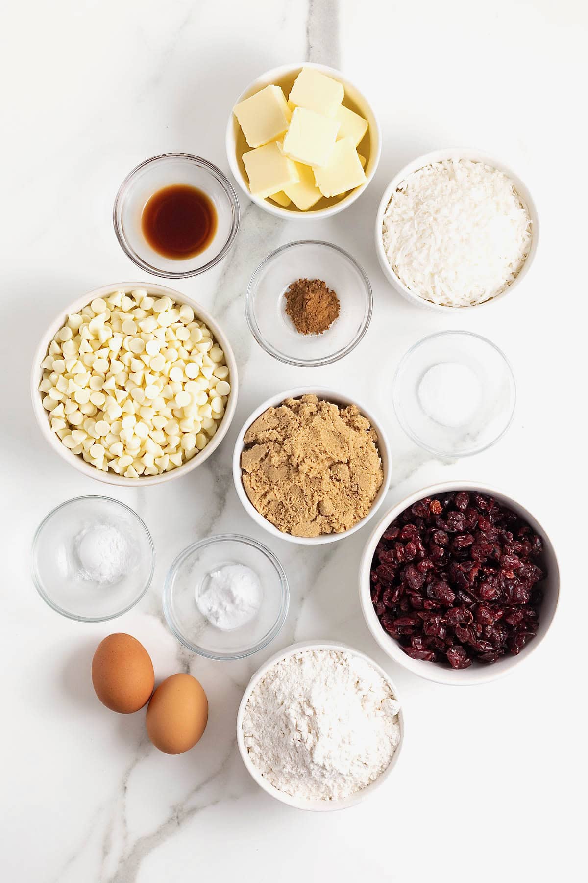 Ingredients to make white chocolate cranberry coconut cookie bars in small glass dishes on a white marble counter.