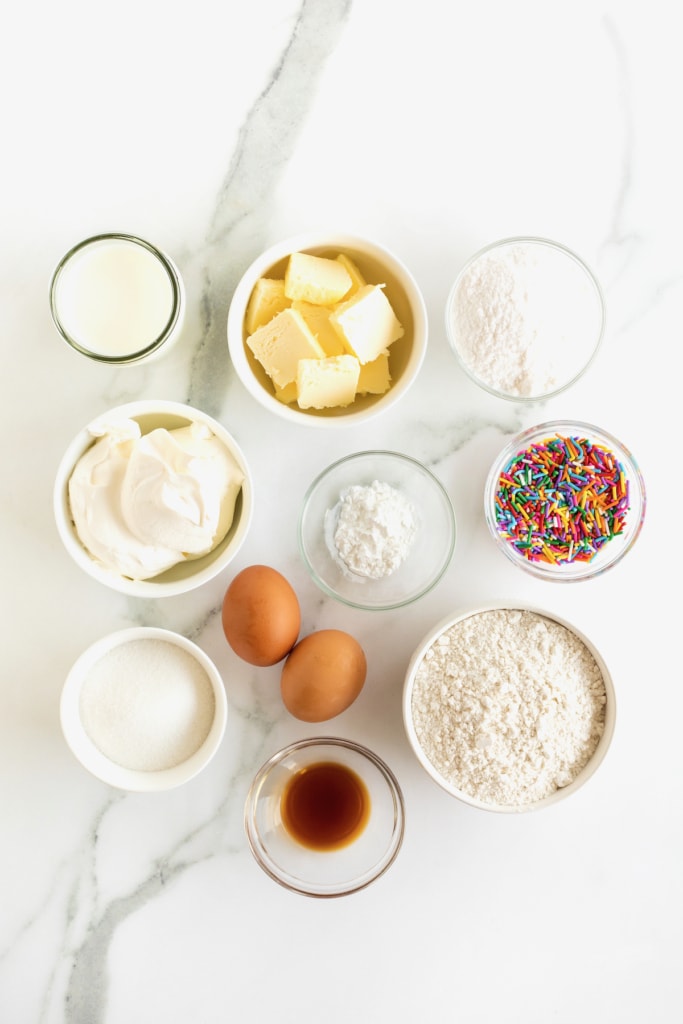 Ingredients for vanilla confetti cupcakes in small white glass dishes on a white marble counter.