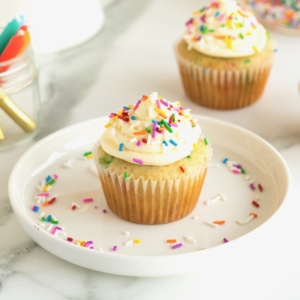 A vanilla cupcake with vanilla frosting and colorful sprinkles on a white plate. Colorful sprinkles are scattered on a the plate. A second cupcake sits in the background on the white marble counter.