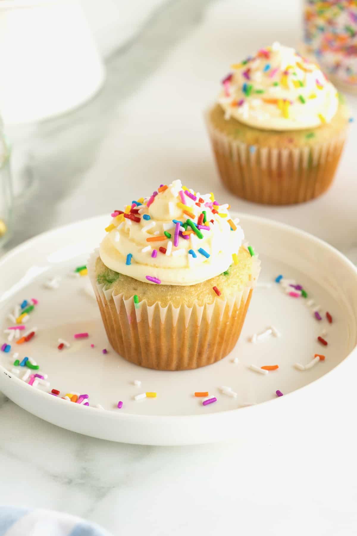 A vanilla cupcake with vanilla frosting and  colorful sprinkles on a white plate. Colorful sprinkles are scattered on a the plate. A second cupcake sits in the background on the white marble counter.