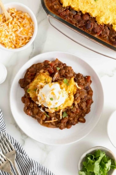 A white dinner plate with a serving of chili cornbread casserole heaped int he middle. The casserole is topped with a dollop of sour cream.