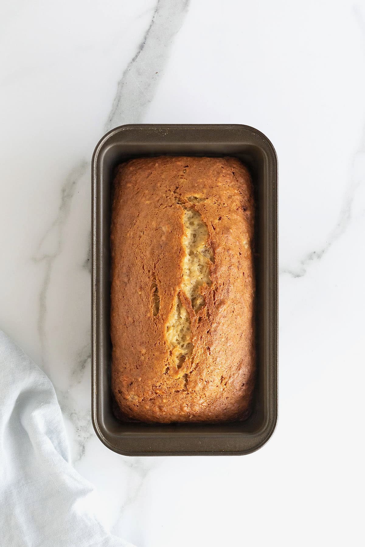 A loaf of brown butter banana bread in a dark aluminum loaf pan on a white marble counter.