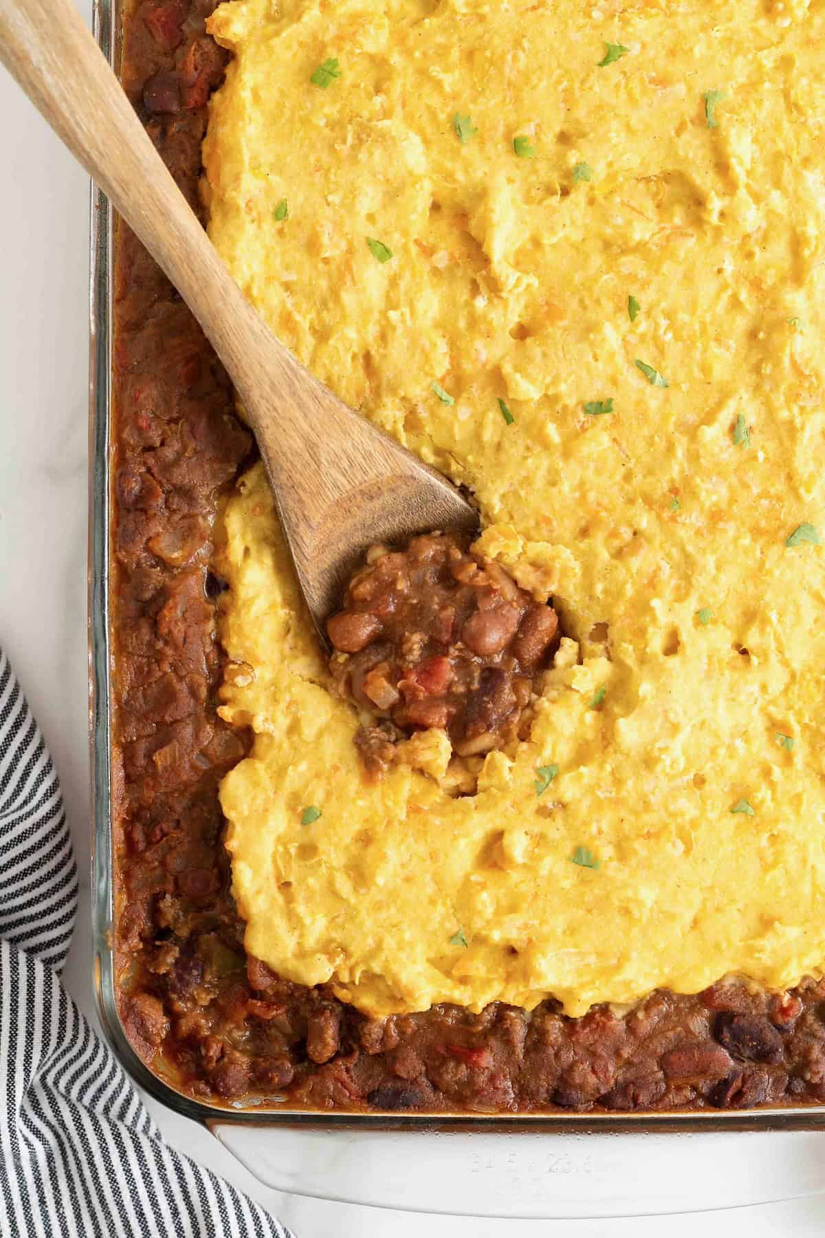 A wooden spoon in a glass baking dish of chili cornbread casserole.