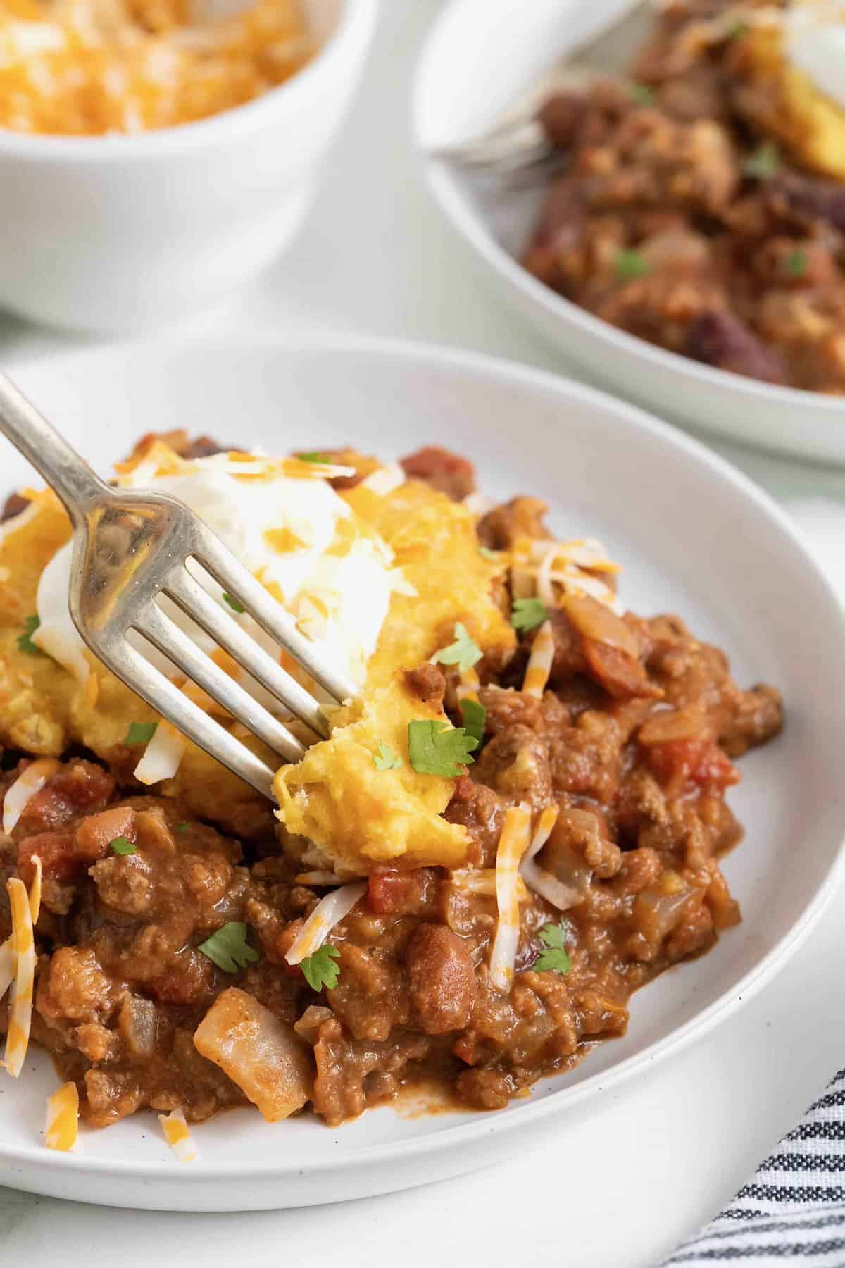 A white dinner plate with a serving of chili cornbread casserole heaped int he middle. The casserole is topped with a dollop of sour cream.