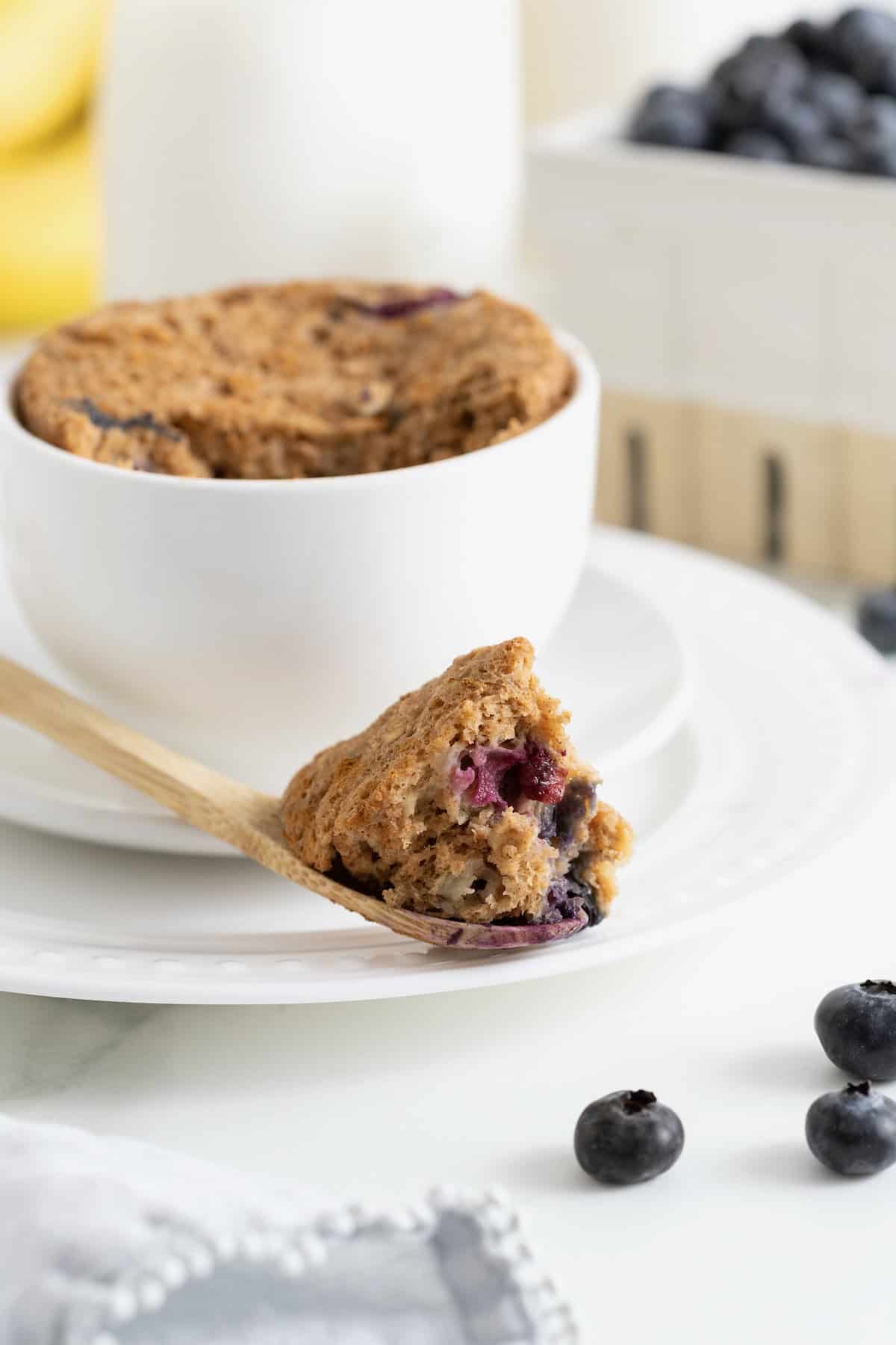 A bite of blueberry banana muffin on a wooden spoon in front of a mug.