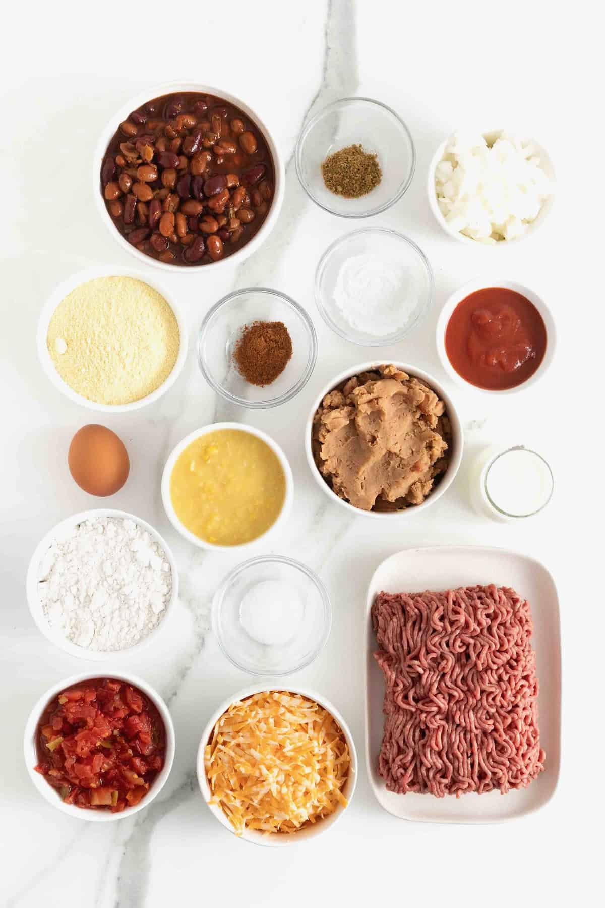 Ingredients for chili cornbread casserole in small glass dishes on a white marble counter.