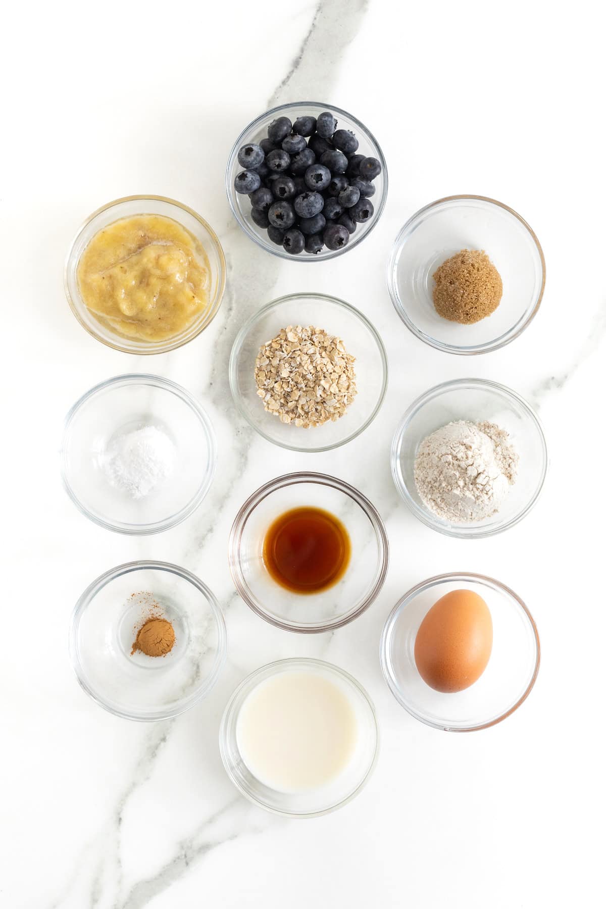 Ingredients to make microwave blueberry banana muffins small glass dishes on a white marble counter.