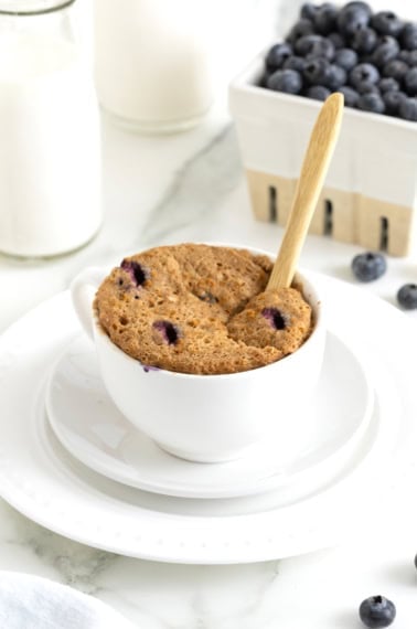 A blueberry banana muffin in a white mug. The mug is sitting on a white saucer.