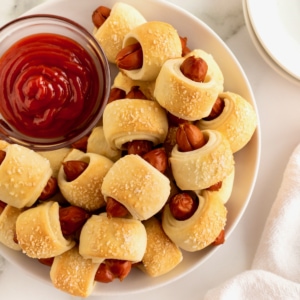 A large white serving plate filled with pigs in a blanket surrounding a small glass dish of ketchup.