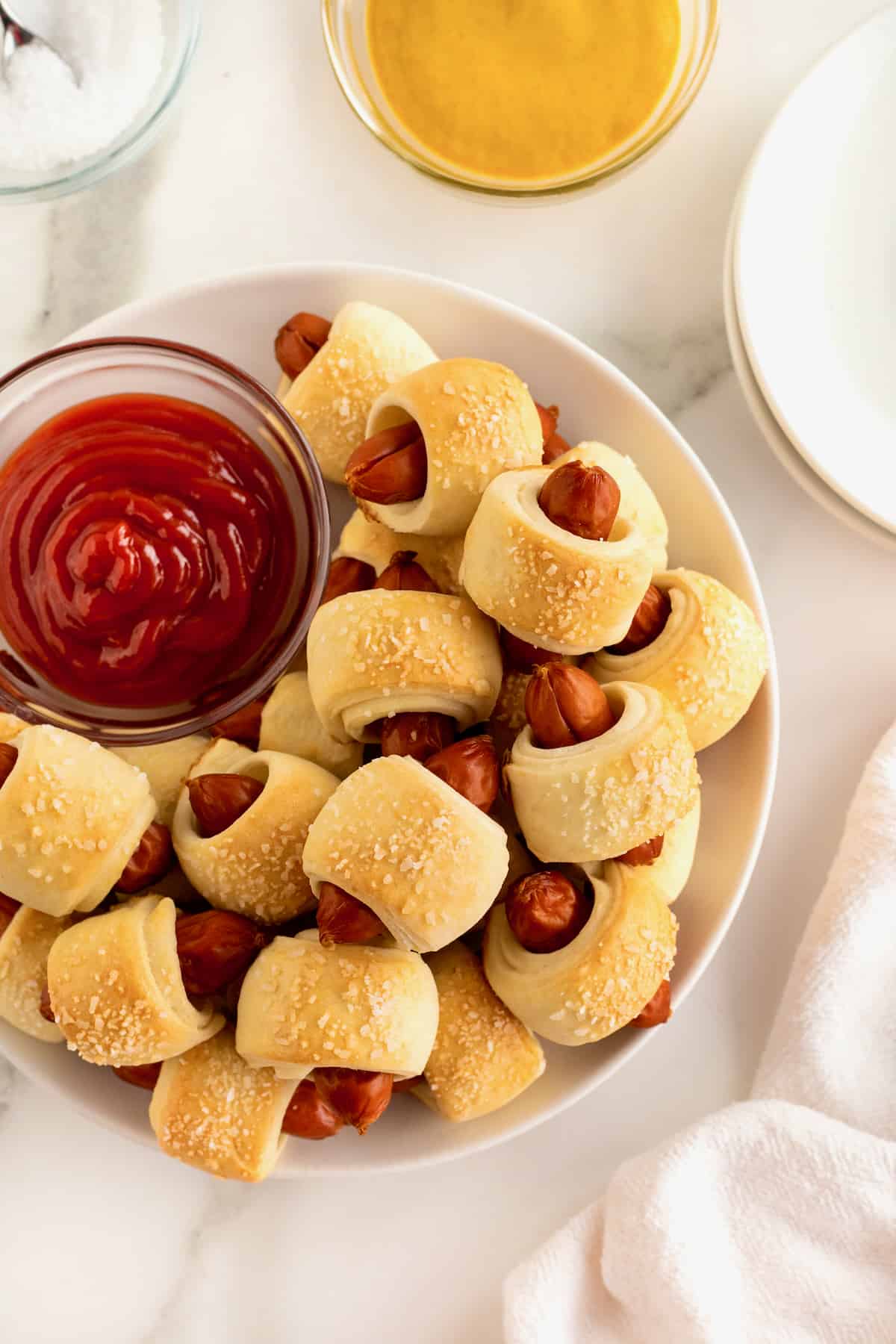 A large white serving plate filled with pigs in a blanket surrounding a small glass dish of ketchup.