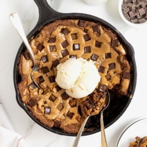 A chocolate chunk cookie in a large black skillet. There's a scoop of ice cream on top and three spoons stuck in it.