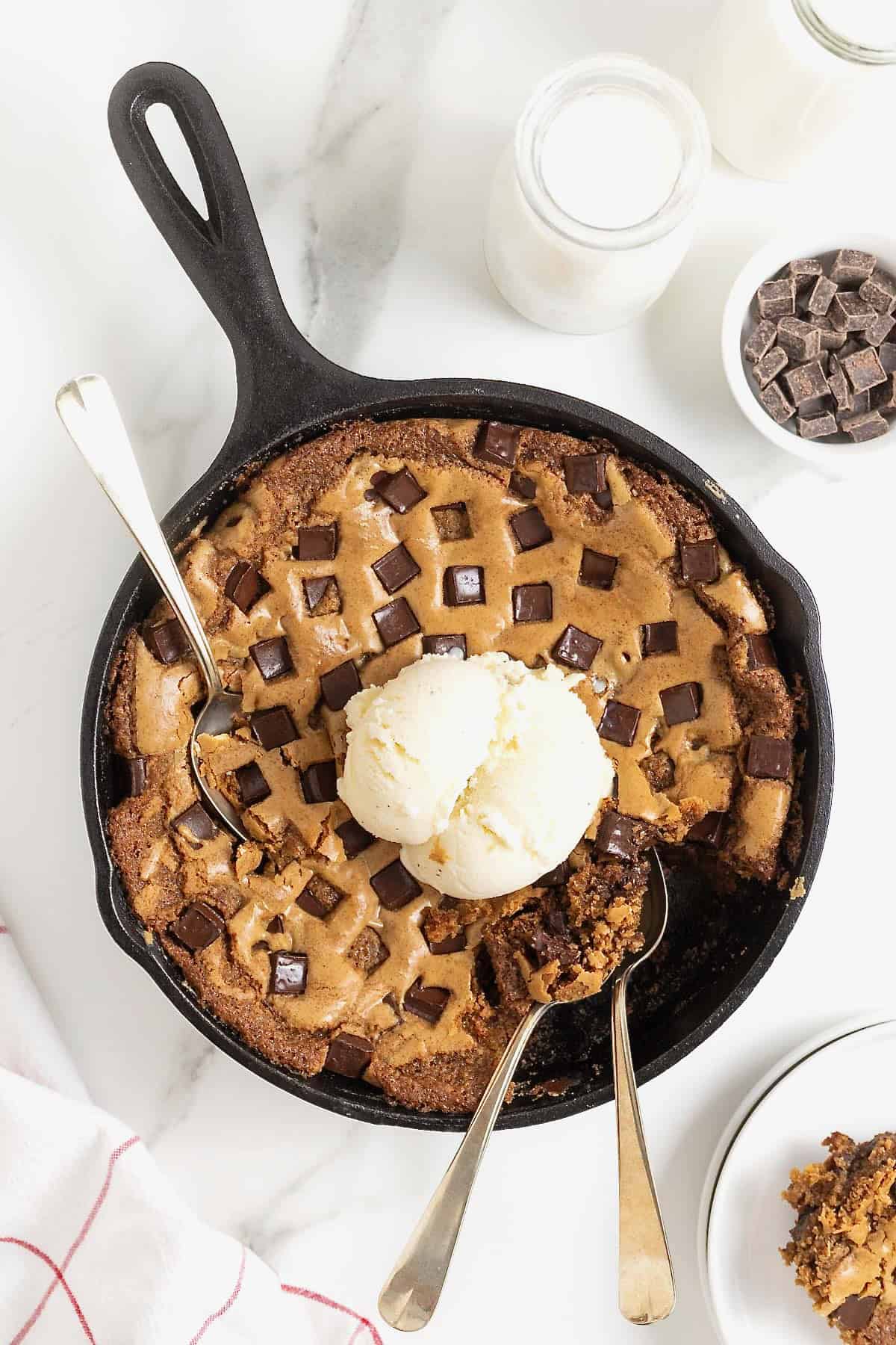 A chocolate chunk cookie in a large black skillet. There's a scoop of ice cream on top and three spoons stuck in it.