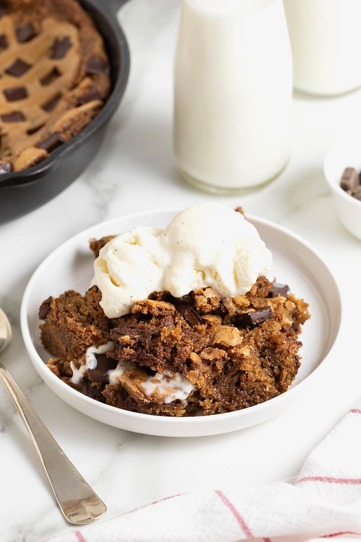 A serving of skillet cookie on a white rimmed plate topped with a scoop of vanilla ice cream.